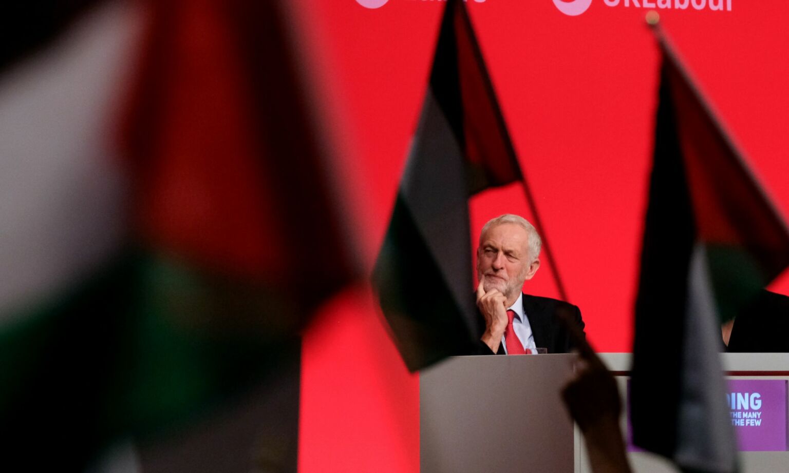 Then Labour leader Jeremy Corbyn sits on stage as delegates wave Palestinian flags at the Labour Party Conference, 25 September 2018.